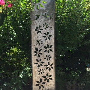 May include: A metal garden sculpture with a cutout design of flowers and a sun. The sculpture is painted in a silver color and has a rustic look.