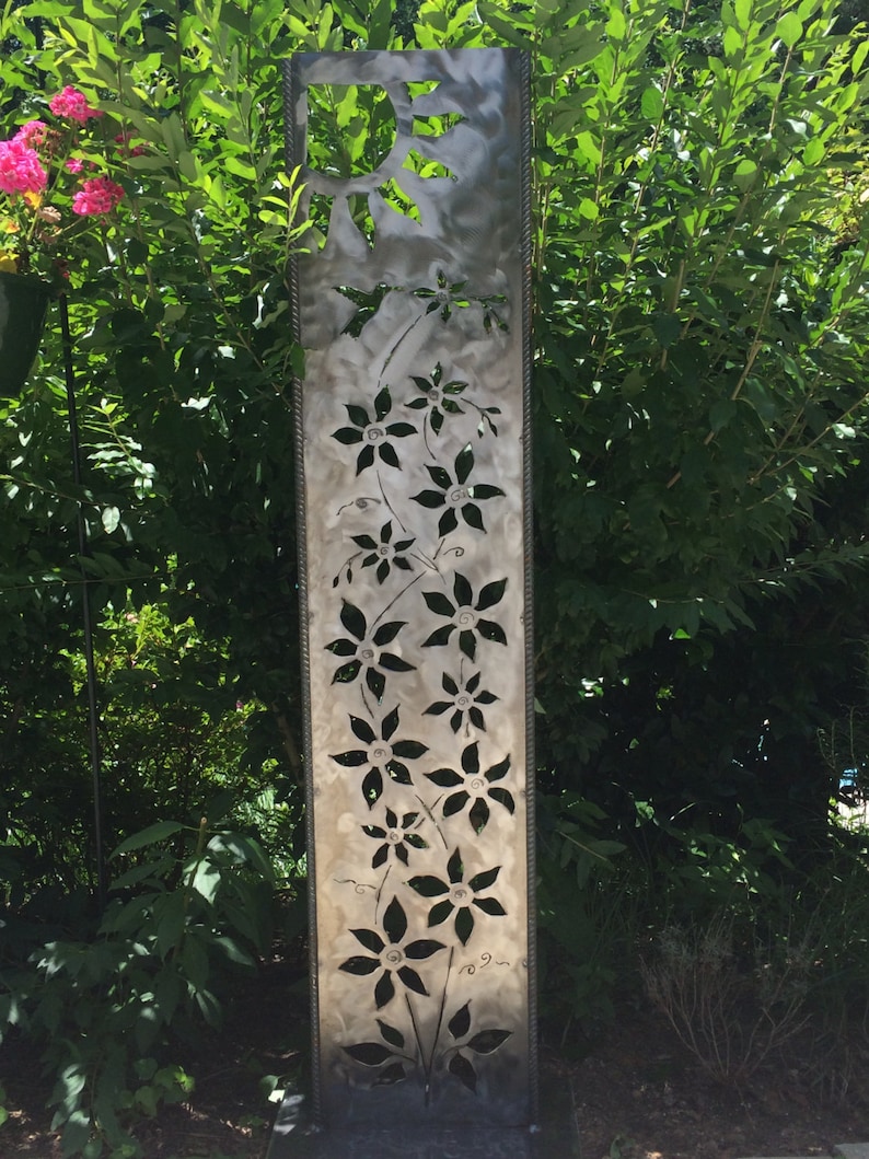 May include: A metal garden sculpture with a cutout design of flowers and a sun. The sculpture is painted in a silver color and has a rustic look.