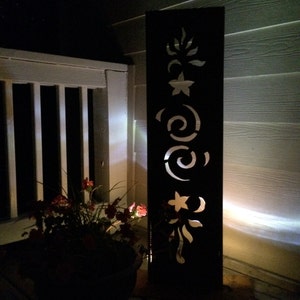 May include: A black metal outdoor light fixture with a decorative floral design cut out. The light is illuminated and casting a warm glow.