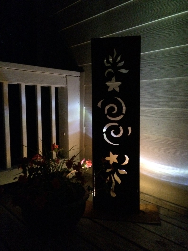 May include: A black metal outdoor light fixture with a decorative floral design cut out. The light is illuminated and casting a warm glow.