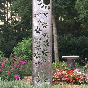 May include: A metal garden sculpture with a cutout design of flowers, a hummingbird, and a sun. The sculpture is in a garden setting with pink and red flowers.