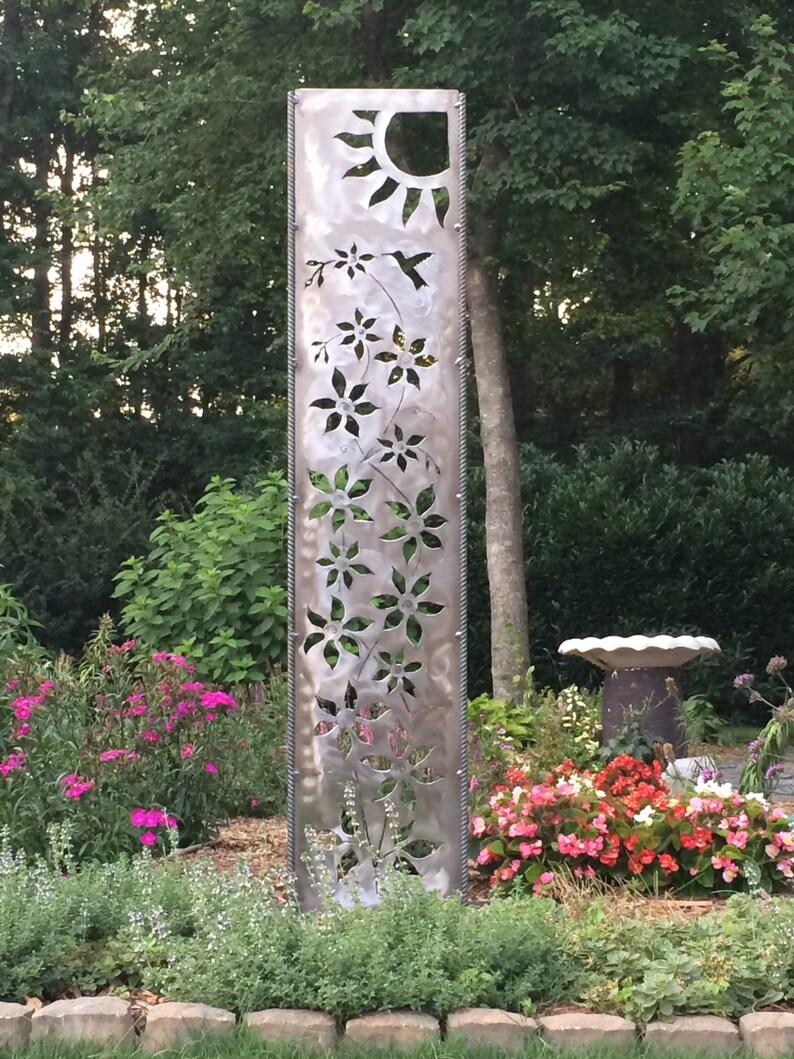 May include: A metal garden sculpture with a cutout design of flowers, a hummingbird, and a sun. The sculpture is in a garden setting with pink and red flowers.