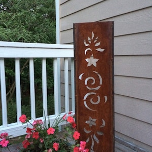 May include: A rusty metal garden sculpture with cut-out designs of stars, swirls, and leaves. The sculpture is standing on a wooden deck next to a potted plant with pink flowers.