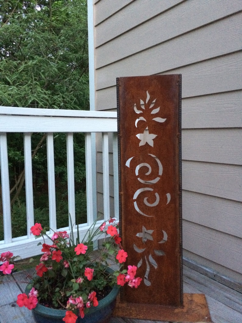 May include: A rusty metal garden sculpture with cut-out designs of stars, swirls, and leaves. The sculpture is standing on a wooden deck next to a potted plant with pink flowers.