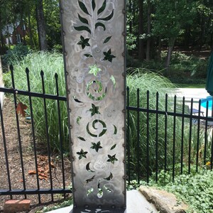 May include: A tall, silver metal garden sculpture with a decorative cut-out design featuring flowers, leaves, and swirls. The sculpture is standing on a concrete patio.