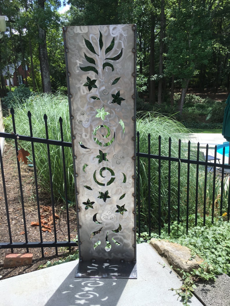 May include: A tall, silver metal garden sculpture with a decorative cut-out design featuring flowers, leaves, and swirls. The sculpture is standing on a concrete patio.