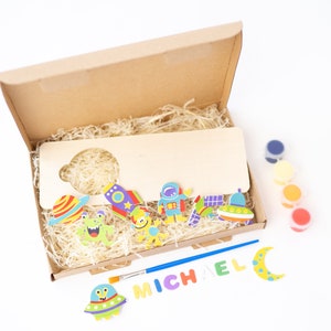 May include: A wooden craft kit with a space theme. The kit includes a wooden sign, paint, a paintbrush, and colourful stickers featuring astronauts, planets, and spaceships. The sign is blank and can be personalised with the name "MICHAEL".