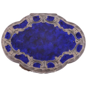 Puede incluir: Una caja de alhajas ovalada de plata con un diseño intrincado alrededor del borde de la tapa. La tapa está decorada con un centro de esmalte azul.