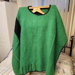 May include: A green poncho with a black accent on the left shoulder and black trim along the neckline and hem. The poncho is displayed on a wooden hanger and has a textured appearance. The garment is made of a thick, warm material.