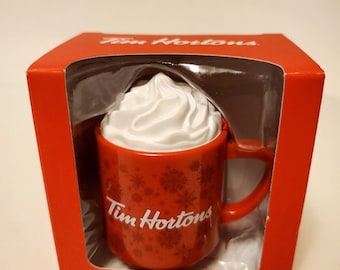 May include: A red Tim Hortons tree ornament in the shape of a coffee mug, topped with white whipped cream. The mug features snowflake patterns and the Tim Hortons logo. It is presented in a red box.