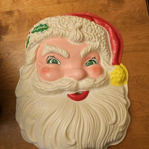 May include: A moulded plastic Santa Claus face wall decoration featuring a white beard, moustache, and textured hat trim. The figure has green eyes, pink cheeks, a red mouth, and a red hat with a yellow pom-pom and green holly leaf detail.
