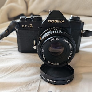 May include: A black Cosina CT-1 35mm film camera with a Cosinon-S 50mm 1:2 lens made in Japan. The camera features a black leatherette body, a top-mounted strap, and a separate 49mm polarising filter cap resting in front of the lens assembly.