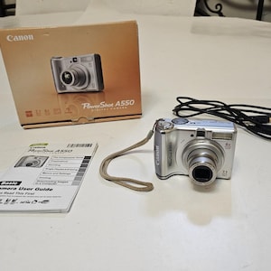 May include: A silver Canon PowerShot A550 digital camera with a wrist strap, a black USB cable, a printed user guide, and the original cardboard box. The camera features a 4x optical zoom lens and 7.1 megapixels, with branding visible on the box.