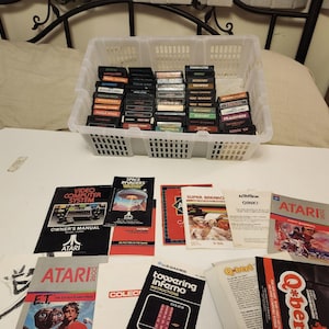 Puede incluir: Una colección de cartuchos y manuales de videojuegos Atari. Los cartuchos están en una cesta de plástico blanco. Varios manuales de juegos están extendidos debajo, incluyendo "Video Computer System" y "Space Invaders".