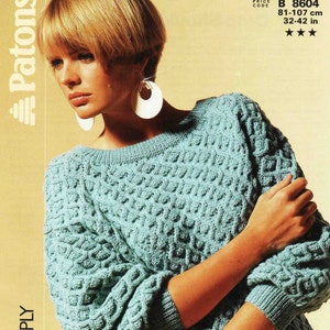 Puede incluir: Un suéter de punto azul verdoso con un patrón de trenza. El suéter se muestra en una modelo con cabello rubio corto. El código de precio es B 8604, 81-107 cm, 32-42 pulgadas.
