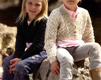 May include: Two young girls are seated on a rock, showcasing children's knitwear. One wears a black cardigan, the other a cream-colored, patterned cardigan. The image includes text that reads "life double knitting" and "Stylecraft". Measurements are listed as 20-30 in.