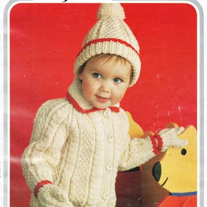 May include: A white knitted cardigan with red trim and buttons, worn by a child with a white knitted hat with a red stripe and a pom-pom. The child is holding a yellow stuffed animal. Hayfield 1496 Aran.