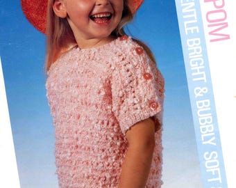 Könnte beinhalten: Ein Mädchen trägt einen rosa Strickpullover mit kurzen Ärmeln und einen Strohhut. Der Pullover hat ein strukturiertes Muster und Knöpfe auf der Schulter. Der Text "POM POM" und "SOFT & GENTLE BRIGHT & BUBBLY SOFT & GENTLE" ist sichtbar.