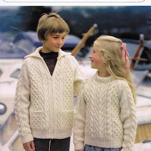May include: Two children wearing white knitted sweaters with cable knit patterns. The boy is wearing a zipped sweater and the girl is wearing a crew neck sweater. The sweaters are designed to fit chest sizes 22-34 inches.