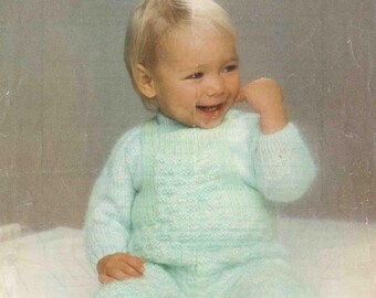 May include: A baby wearing a light blue knitted sweater and pants set. The image has text that reads "Patons FAIRYTALE Misty DK" and measurements of 18-20 inches and 46-51 cm. The baby is smiling and sitting on a white surface.