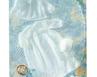 May include: A white baby outfit set, including a dress, jacket, and bonnet, displayed on a blue and white patterned blanket. The set is made of a delicate, possibly knitted, fabric. The Patons logo is visible at the top.