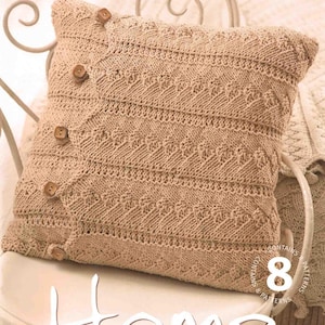 May include: A beige knitted pillow with a buttoned closure, resting on a white chair. The pillow features a textured pattern and is part of a home decor collection. The word "Home" is visible in the image.