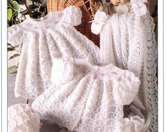 May include: A collection of white, knitted baby clothes, including dresses, a bonnet, booties, and a blanket. The dresses have short sleeves and are adorned with small pink flowers. The image also includes a doll in a wicker basket. The text "BABY 3 PLY" is visible.