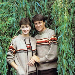 May include: A couple wearing matching knit sweaters with geometric patterns in shades of brown, red, and white. The sweaters have a crew neck and long sleeves. The couple is standing in front of a green leafy background.