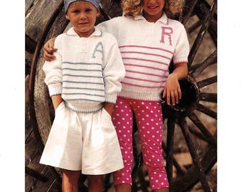 May include: Two children wearing knitted outfits. One child wears a white sweater with blue stripes and the letter "A", white shorts, and a blue bandana. The other child wears a white sweater with pink stripes and the letter "R", pink polka dot pants, and a white beret. The image includes the text "Patons" and "Double Knitting".