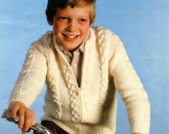 Könnte beinhalten: Ein Vintage-Strickmuster-Cover mit einem lächelnden Kind in einem cremefarbenen Zopfmuster-Reißverschluss-Pullover. Der Pullover hat ein strukturiertes Design und wird mit einer braunen Cordhose kombiniert. Das Sirdar-Logo ist sichtbar.