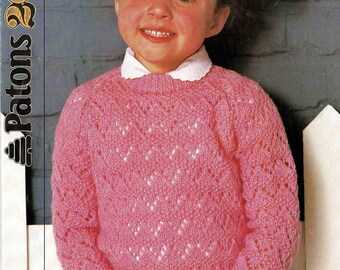 May include: A young girl wearing a pink knitted sweater with a white collar. The sweater has a textured, openwork design. The image also includes text that reads "Patons 200 DK" and measurements of 20-30 inches.