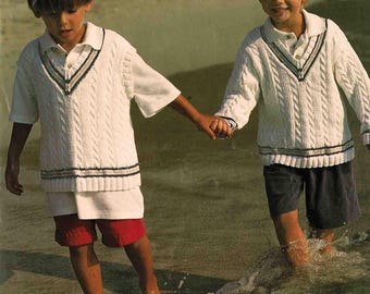 May include: Two young children wearing white cable-knit sweaters with navy and green trim, paired with shorts, walk hand-in-hand through shallow water. The image features the text "Patons" and sizing information for children aged 2-11 years.
