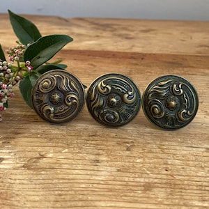 May include: Three antique brass cabinet knobs with ornate floral designs. The knobs are round and have a dark green patina.