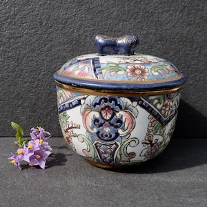 Puede incluir: Un pequeño tarro redondo de cerámica con tapa. El tarro está decorado con un colorido patrón floral en tonos azules, rosas y verdes. La tapa tiene un pequeño pomo en forma de pájaro.