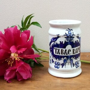 Può includere: Un vaso di ceramica bianca con disegni floreali blu e bianchi e il testo "TABAC RAPPE" sul davanti. Il vaso è appoggiato su una superficie di legno con un fiore di peonia rosa in primo piano.