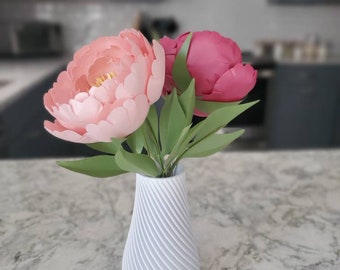 May include: A white 3D-printed vase with a spiral texture holds two large paper flowers. One flower is light pink with yellow centers, and the other is dark pink. Both flowers have green paper leaves and stems extending from the vase.