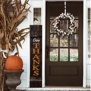 May include: A wooden sign with the words "Give Thanks" in orange letters on a dark brown background. The sign is leaning against a white door with a cotton wreath hanging on it.