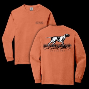 May include: A long-sleeved, orange t-shirt with a black and white graphic of a hunting dog on the back. The text "Wild Whiskey Apparel Co. Outdoors" is printed below the dog. The front of the shirt has a small logo that says "Wild Whiskey Apparel Co. Outdoors".