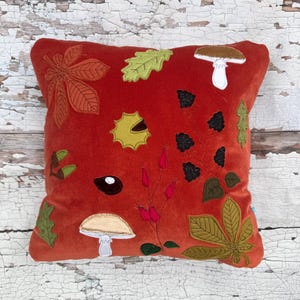 May include: A square rust-orange velvet throw pillow decorated with embroidered nature motifs including brown chestnut leaves, green oak leaves, white mushrooms, black blackberries, pink rose hips, acorns, and a spiky yellow chestnut husk.