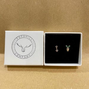 May include: A white box with the text "TUTSHILL COMPANY" and a logo of antlers. Inside, a pair of small earrings: one shaped like a shovel, the other a potted plant with green leaves. The earrings are on a black background.