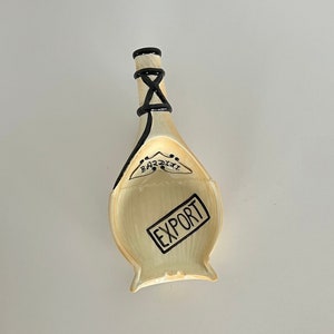May include: A ceramic bottle stopper shaped like a wine bottle with black and white details. The stopper has the words "EXPORT" and "BAZZINI" printed on it.