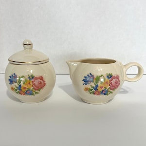 Könnte beinhalten: Ein vintage cremefarbenes Zucker- und Sahne-Set mit einem floralen Kreuzstich-Design. Das Set besteht aus einer runden Zuckerdose mit Deckel und einem Sahnekännchen mit Henkel.