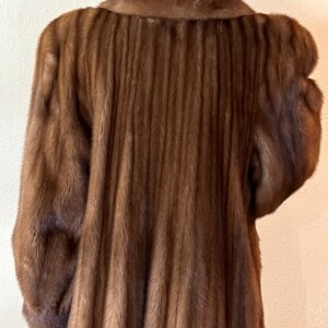 May include: A long brown fur coat with a vertical ribbed pattern. The coat has a classic collar and long sleeves.