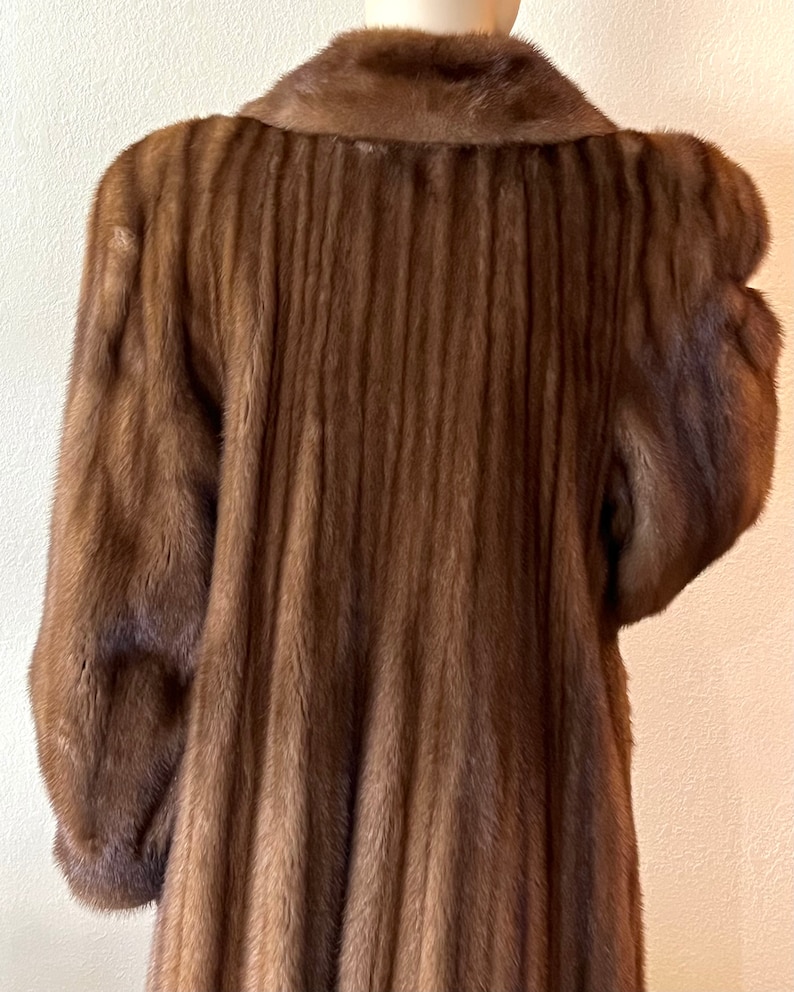 May include: A long brown fur coat with a vertical ribbed pattern. The coat has a classic collar and long sleeves.