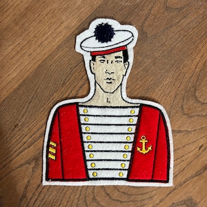May include: Embroidered patch featuring a sailor in a red jacket with gold buttons and an anchor emblem. The sailor wears a white hat with a black pom-pom. The patch has a white border and is set against a wooden background.