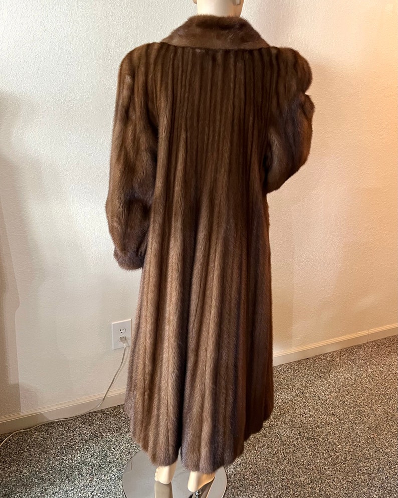 May include: A long brown fur coat with a collar and a pleated design. The coat is on a mannequin.