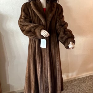 May include: A long brown fur coat with a large collar. The coat is on a mannequin and has a white tag hanging from it.