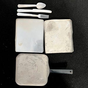以下が含まれることがあります： スプーン、ナイフ、フォーク、2つの長方形の食品容器、黒いハンドル付きの正方形のフライパンなど、ヴィンテージの銀色のメスキットアイテムのコレクション。アイテムは暗い表面に配置されています。