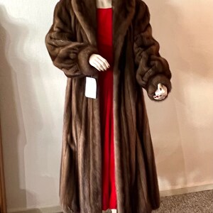 May include: A long brown fur coat with a large collar and long sleeves. The coat is being worn by a mannequin and is displayed against a white wall.