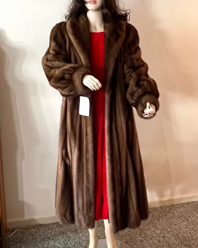 May include: A long brown fur coat with a large collar and long sleeves. The coat is being worn by a mannequin and is displayed against a white wall.